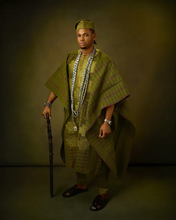 A man in an olive and emerald patterned agbada holding a carved walking stick