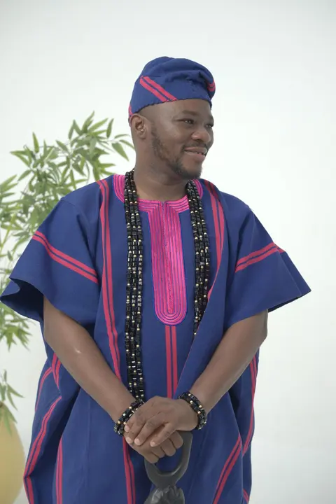 A man in an indigo aso oke agbada with magenta stripes and a matching cap