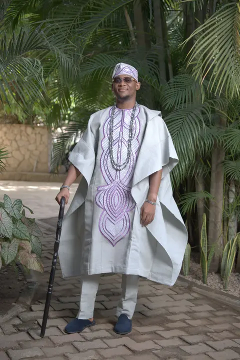A man in a white agbada with lilac embroidery, standing among palms with a cane