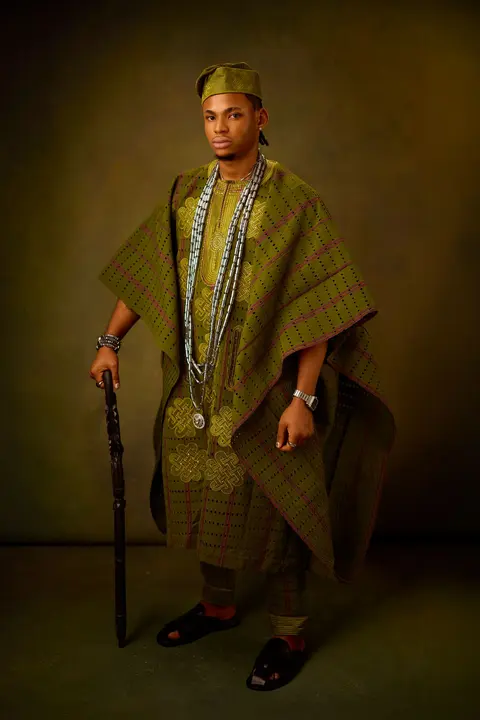 A man in an olive check aso oke agbada holding a carved staff