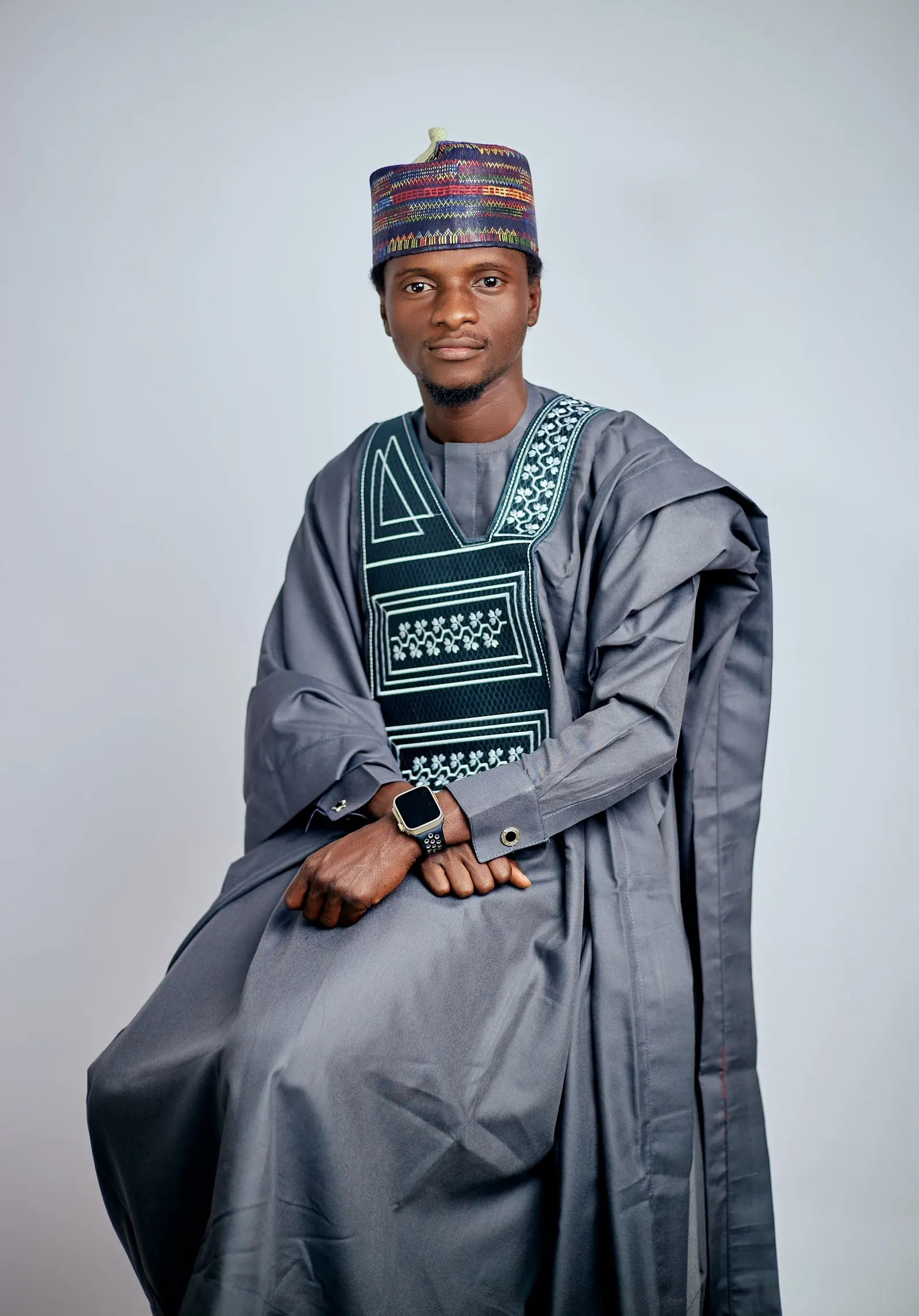 A man seated in a grey embroidered agbada and patterned cap against a pale backdrop